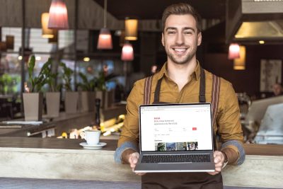 handsome young waiter holding laptop with airbnb website on screen in cafe