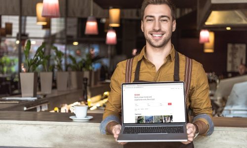 handsome young waiter holding laptop with airbnb website on screen in cafe
