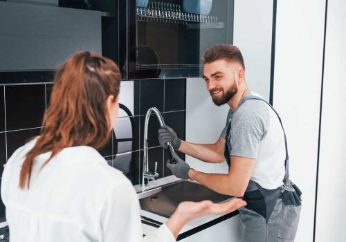 Young professional plumber in grey uniform shows results of work for housewife on the kitchen Young professional plumber in grey uniform shows results of work for housewife on the kitchen